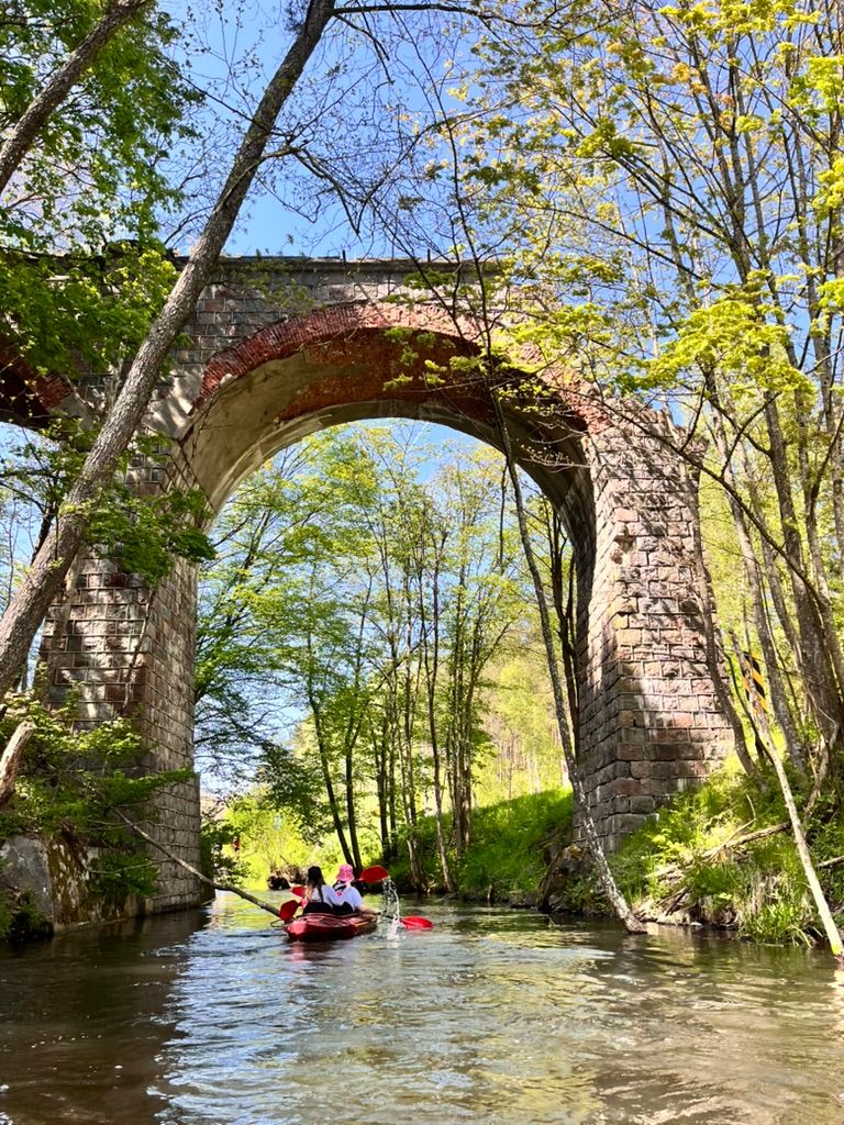 Dwa kajaki płyną pod łukiem starego wiaduktu kolejowego na rzece Łupawa