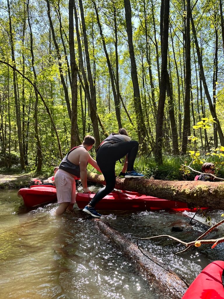 Uczestnicy wspinają się po kłodzie nad rzeką, kajaki w tle